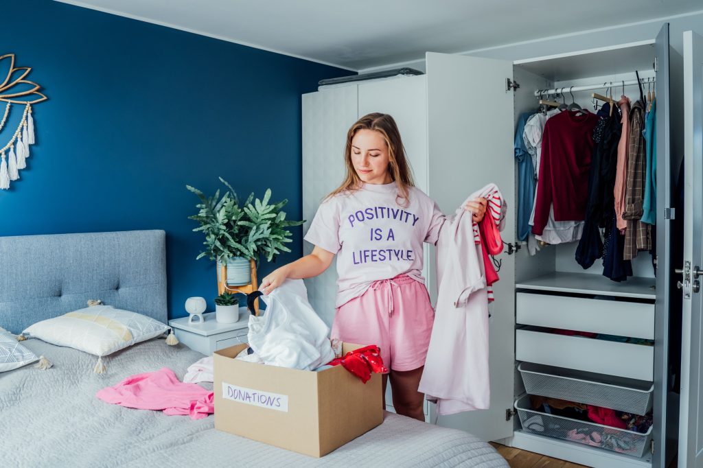 Woman putting selected clothes from her wardrobe for donating to a Charity shop. Decluttering, Sorting clothes and Cleaning Up. Reuse, second-hand concept. Conscious consumer, sustainable lifestyle.